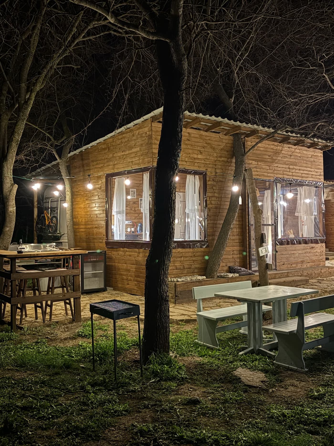 Обща градина на Elysium вечер — маса за хранене на открито, барбекю и топли светлини между дърветата